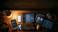 Modern workspace setup with multiple devices showing coding interfaces and development tools on wooden desk with coffee cup and headphones.