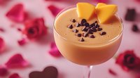 Creamy chocolate cocktail garnished with fresh mango slices and chocolate chips in coupe glass, romantic pink rose petal background for Valentine's Day celebration.