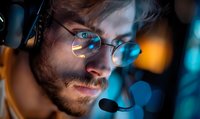 Young bearded Caucasian male wearing glasses and gaming headset in dark room with blue and orange ambient lighting, close-up profile view showing focused expression.