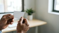 Senior male with dark skin tone holding blank white card in modern office setting with wooden desk and potted plant, suitable for business, corporate messaging, and professional communications.