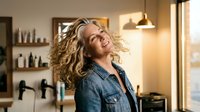 Happy blonde woman with curly hair dancing in modern kitchen wearing denim shirt expressing joy and freedom in warm sunlit home interior.