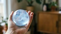 Hand holding transparent crystal ball with blue reflections in cozy home interior with plants and warm lighting for fortune telling and mystical practices.