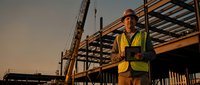 Male construction worker in safety vest holding tablet at building site during golden hour sunset with steel framework and scaffolding in background.