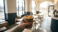 Hand holding coffee cup with latte art in modern cafe interior with arched windows and warm natural lighting for hospitality and lifestyle concepts.