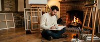 Young man artist painting on canvas while sitting on floor near fireplace in cozy studio workspace with easels and warm lighting atmosphere.