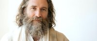 Middle-aged Caucasian man with long wavy hair and full beard wearing white robe, serene expression for spiritual wellness meditation concepts.