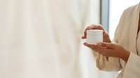 Hands holding white cosmetic jar with cream or skincare product against neutral beige background for beauty and wellness marketing.