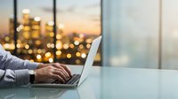 Businessman working on laptop at desk with city skyline and golden sunset lights in background, remote work and urban business concept.