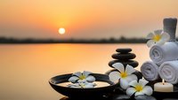 Spa wellness setup with plumeria flowers, stacked stones, towels and candles at golden sunset by water for relaxation and meditation treatments.