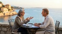 Senior couple enjoying romantic dinner conversation at seaside restaurant terrace during golden hour sunset with Mediterranean coastal village backdrop.
