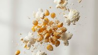 Popcorn kernels and fluffy white popped corn exploding in mid-air against neutral background, perfect for snack food advertising and cinema concepts.