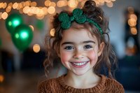 Young girl with curly brown hair wearing green glitter shamrock hair bow smiles brightly against festive bokeh background with green balloons, celebrating St Patrick's Day.