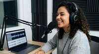 Young mixed-race woman podcaster recording in professional studio with microphone, headphones and laptop showing audio waveforms for broadcasting content.