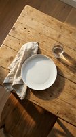 Overhead view of empty white ceramic plate with linen napkin and water glass on rustic wooden table for dining, restaurant, or food styling projects.