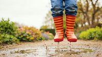 Child jumping in puddle wearing bright red and orange striped rain boots, water splashing on wet path with colorful garden background.
