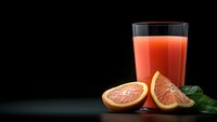 Fresh grapefruit juice in tall glass with cut pink grapefruit halves and green leaves on dark background, healthy citrus drink with copy space.