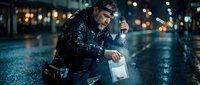 Middle-aged Caucasian man in leather jacket kneeling on wet city street at night, holding lantern with warm light creating atmospheric urban scene with bokeh background.