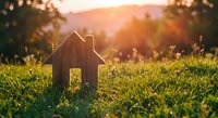 Wooden house model on green grass at sunset with warm golden light and bokeh background for real estate and home ownership concepts.