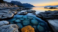 Dramatic rocky coastline with turquoise water pools at sunset, snow-covered stones creating natural patterns against mountainous backdrop and golden sky.