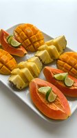 Fresh tropical fruit platter with sliced mango, pineapple and papaya garnished with lime wedges on white serving tray for healthy eating and summer dining.
