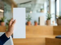 Hand holding blank white paper in modern office with plants and wooden desk, suitable for mockups, advertising, and business presentations.