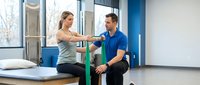 Young blonde woman doing resistance band exercise with male trainer in modern gym rehabilitation center for strength training and physical therapy.