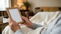Senior adult with aged hands holding blank wooden frame while resting on bed with white bedding, suitable for home decor mockups, lifestyle photography, and interior design presentations.