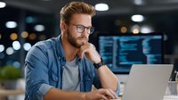 Young Caucasian male programmer with glasses working on laptop in modern office environment with coding screens in background for tech projects.