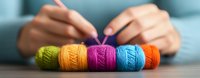 Colorful yarn balls arranged in row on wooden table with blurred hands holding knitting needles in background, creating cozy crafting atmosphere.