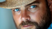 Close up portrait of bearded man with intense blue eyes wearing straw hat against blurred background, dramatic lighting highlights facial features and expression.