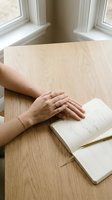 Woman hands resting on wooden table next to open book by window. Peaceful reading moment in natural light for relaxation and mindfulness concepts.