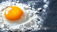 Raw egg yolk on scattered white flour against dark slate background, macro food photography with selective focus creating dramatic culinary composition.