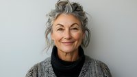 Confident mature Caucasian woman with gray hair and natural makeup wearing turtleneck and cardigan smiling warmly against light background. For lifestyle and aging concepts.