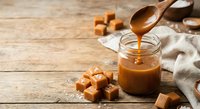 Homemade salted caramel sauce being poured from wooden spoon into glass jar with caramel candies on rustic wooden background for dessert recipes.