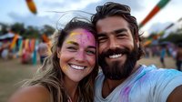 Joyful couple covered in colorful powder paint taking selfie at outdoor festival celebration with blurred festive decorations in background.