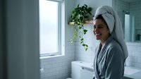Young Caucasian woman in gray robe and towel turban smiling in modern white bathroom after shower, fresh and relaxed morning routine.