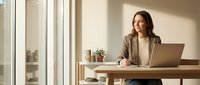 Professional businesswoman working on laptop at modern office desk with natural sunlight streaming through windows creating warm atmosphere for productivity.