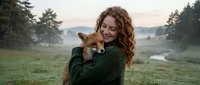Young woman with curly red hair holding adorable fox cub in peaceful countryside setting during golden hour sunset for wildlife conservation concepts.