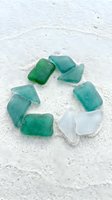 Polished sea glass pieces in green, turquoise, and white arranged in circle on sandy beach. Frosted gems for coastal decor, jewelry making, crafts.