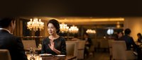 Professional Asian businesswoman dining in upscale restaurant with elegant chandelier lighting, enjoying fine dining experience during business meeting.