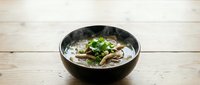 Asian noodle soup with mushrooms and fresh herbs in black bowl on wooden table background for restaurant menu and food photography.