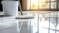 Cleaning white glossy floor with mop and bucket in sunlit room. Shiny reflective surface shows professional maintenance of modern interior space.