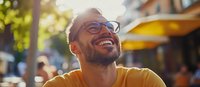 Young Hispanic man with glasses wearing yellow t-shirt laughing outdoors on sunny day, showing genuine joy and happiness in casual urban setting. Natural lifestyle portrait.