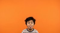 Young boy with dark hair and medium brown skin tone wearing striped shirt, sticking tongue out playfully against vibrant orange background for children's content.