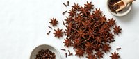 Star anise and cloves scattered on a white surface with small ceramic bowls, aromatic spices for cooking, baking, and herbal tea recipes.