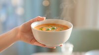 Hot steaming vegetable soup in white bowl held by hands with colorful ingredients and herbs for comfort food dining and healthy eating lifestyle.