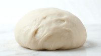 Smooth, rounded ball of raw dough on white surface, ready for baking or rising. Use for bakery, cooking, and food preparation content.