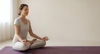 Young Asian woman practicing meditation in lotus pose on purple yoga mat at home, peaceful mindfulness exercise for wellness and mental health relaxation.