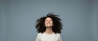 Young mixed-race woman with curly hair laughing joyfully against gray background. Authentic happiness and positive emotions for lifestyle content.