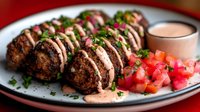 Grilled meat patties with creamy sauce drizzle and fresh herbs served with diced tomatoes and tahini dip on white plate for Mediterranean cuisine presentation.